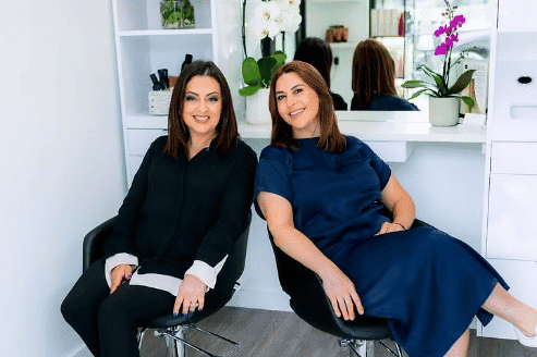 Two women smiling and seated in a modern, well-lit salon with white walls and stylish decor.